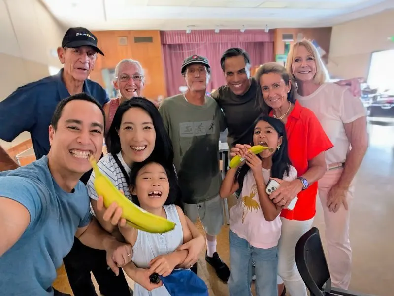 A group of smiling volunteers of different ages taking a selfie in a community hall, one holding a banana.