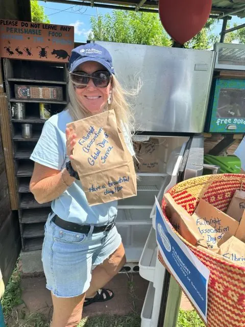 A volunteer in a Fulbright Association cap holding a paper bag marked 'Fulbright Association Global Day of Service. Have a beautiful day!' beside a community food pantry.